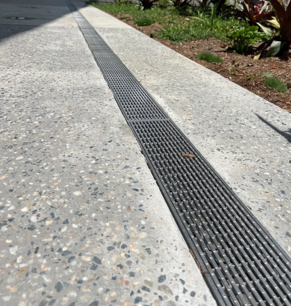 Close-up view of a stormwater drain on a concrete sidewalk, with a parked truck in the background. The drain is flanked by a small landscaped area with plants. Urban setting with buildings and trees under a cloudy sky.