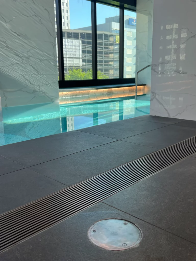 Indoor swimming pool with modern design, featuring large windows that offer a view of city buildings. The pool area has dark tiled flooring and sleek finishes with a visible drain near the edge. Natural light illuminates the water.