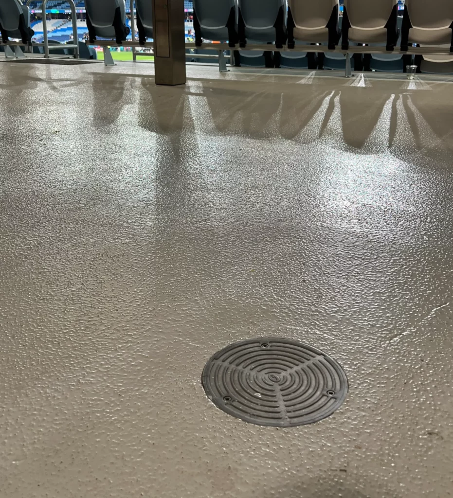 A stadium interior with rows of empty seats and a football field in the background. The smooth floor in the foreground has a drainage cover. The view overlooks the field, with the stadium lights creating reflections on the floor.