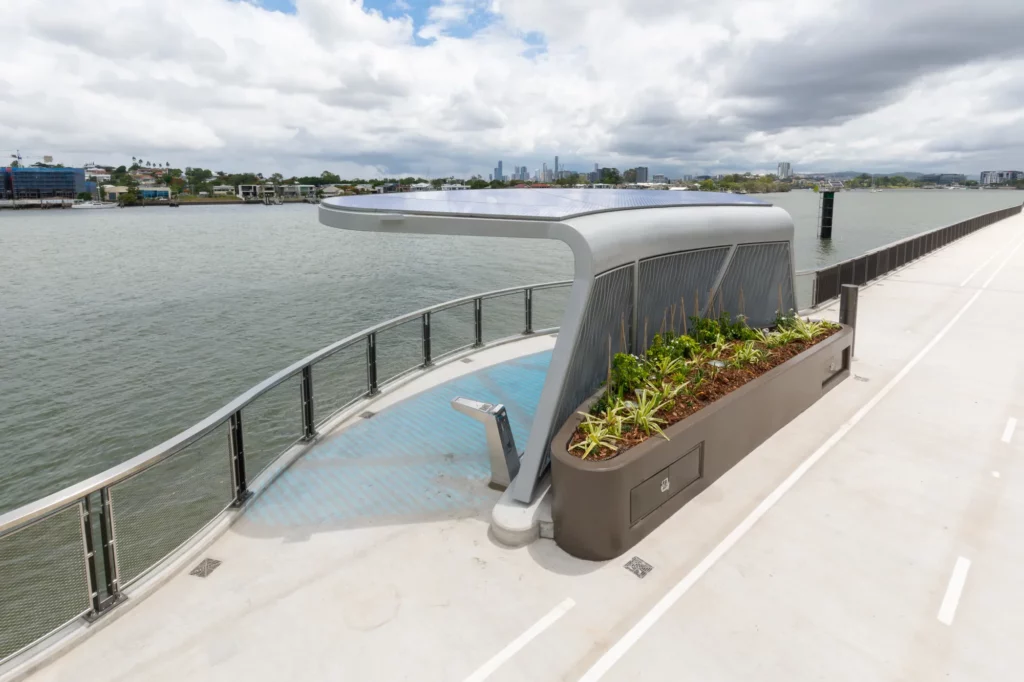 A modern riverfront walkway with a sleek, sculptural viewing platform featuring a planter with greenery. The river is on one side, and a cloudy sky and distant cityscape are visible in the background.