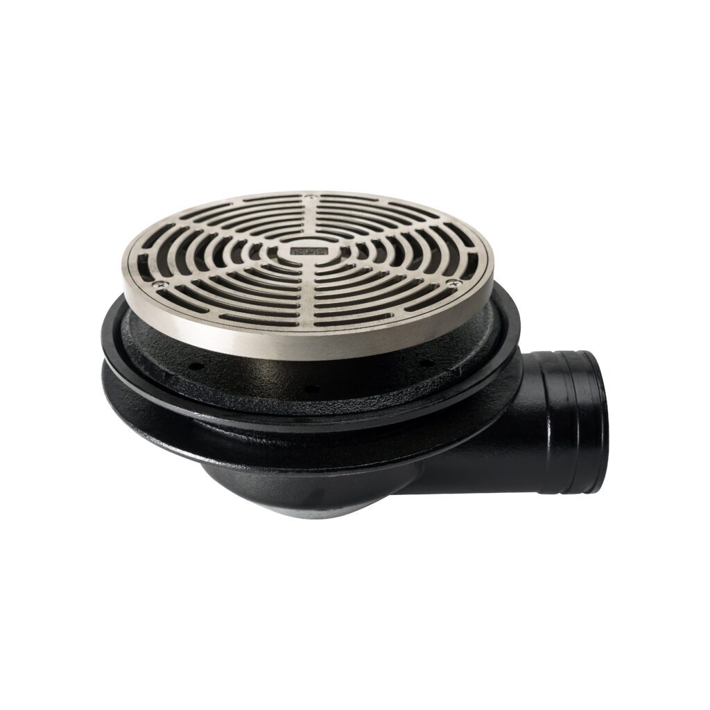 A metal floor drain featuring a round, slotted cover on top and a black horizontal pipe extending to the right. The cover has a grid-like pattern, and the overall design is industrial. The background is plain white.