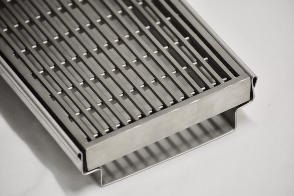 Close-up of a rectangular stainless steel grate with parallel bars and a solid frame, placed on a white surface. The metal grate appears durable and industrial in design.